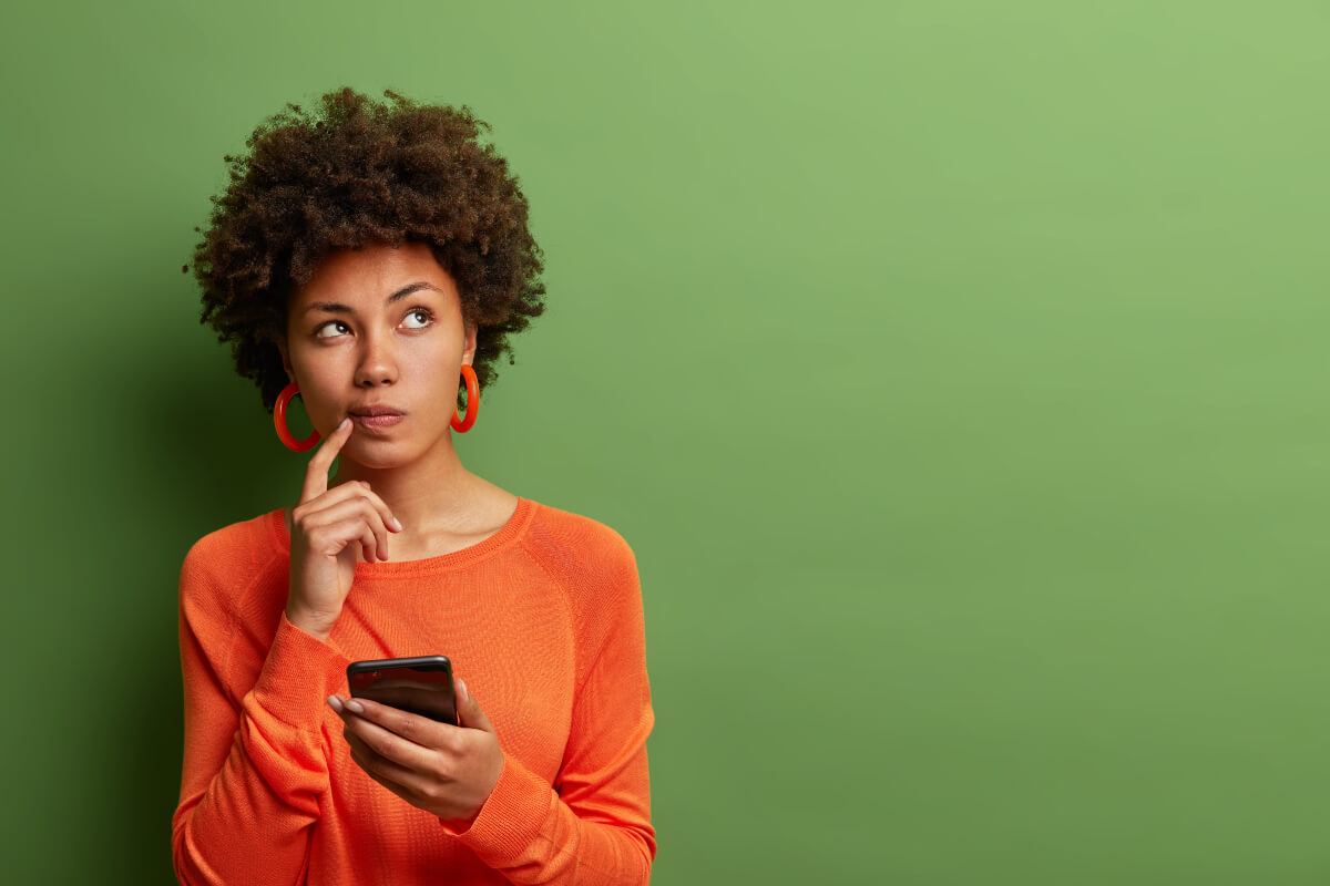 Femme sur fond vert, doigt sur la bouche, smartphone à la main, se demande qui choisir pour son forfait mobile YouPrice entre le réseau d'Orange ou celui de SFR
